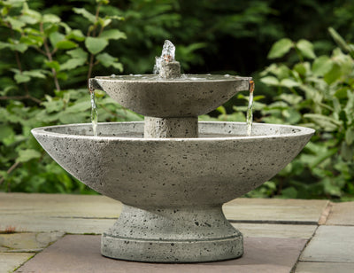Oval shaped two tiered fountain  with two cupper spouts in either sides spilling water in lower bowl,  pictured on grey stones in front of shrubs