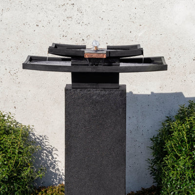 Black Asia-inspired fountain with rectangular bowls and flat copper spout on square pedestal, pictured against white wall
