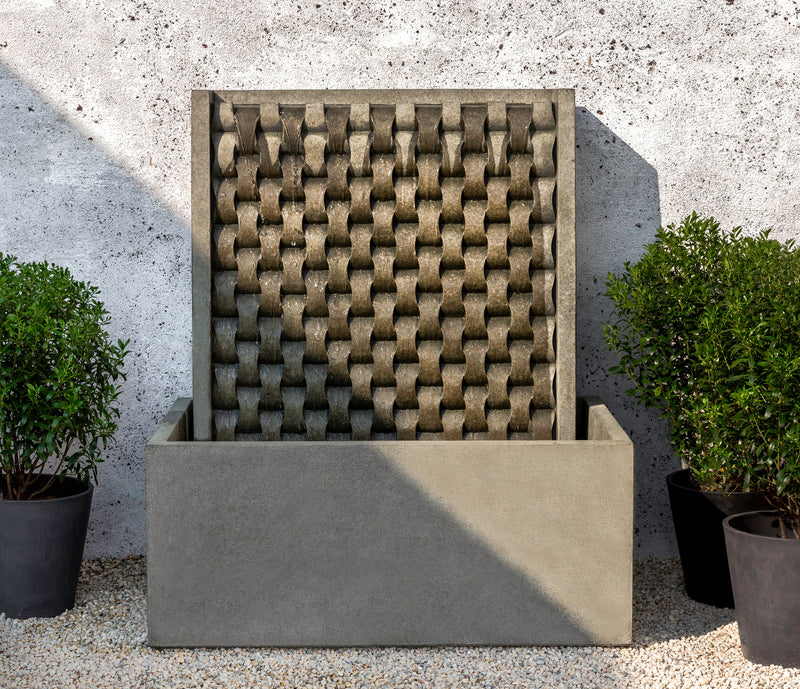 Wall fountain in grey finish with weaved back pictured with water flowing down and potted containers next to it