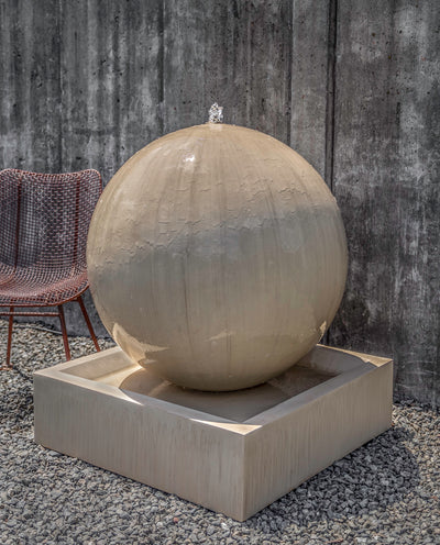 Large spherical fountain on a square base, pictured in front of a grey wall and with a red chair