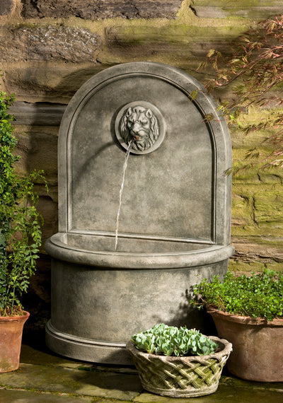 Grey wall fountain with curved back and lion faced medallion , displayed with planted containers 