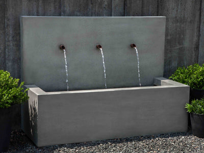 Long rectangular fountain with three spouts pictured against a grey wall