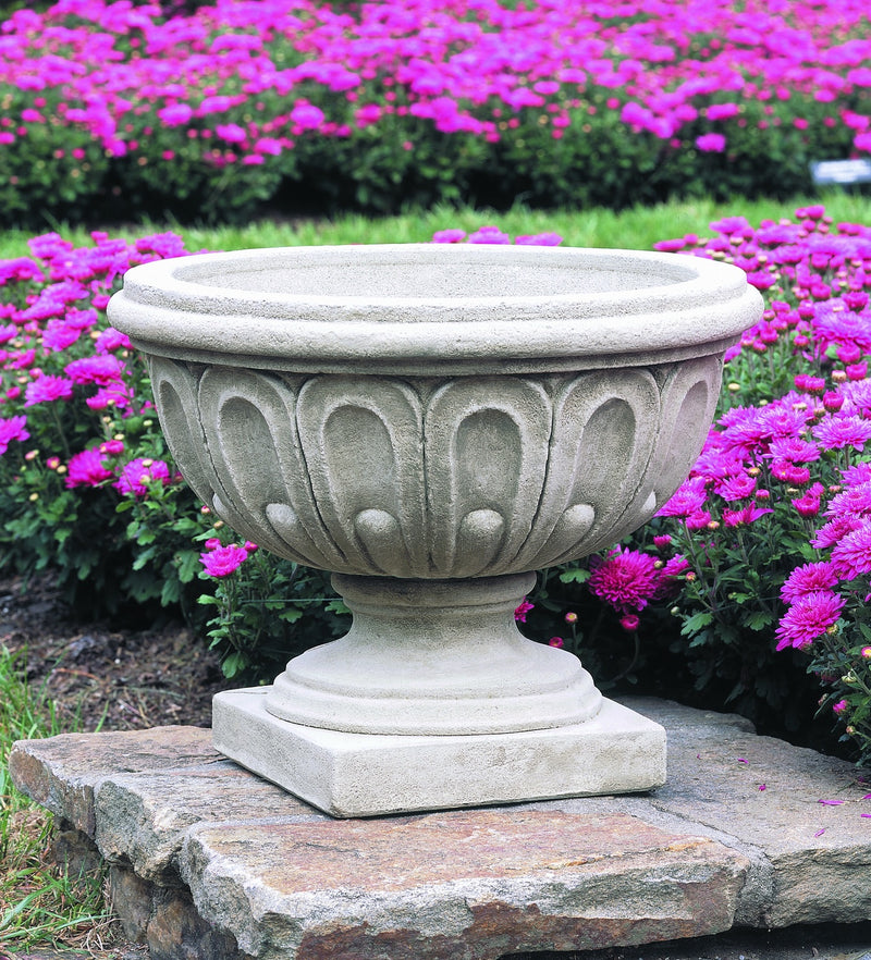 Fluted urn shown on a stone plinth in front of purple mums