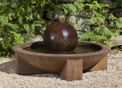 Brown bowl with central sphere spilling water through copper spout displayed on gravel and ferns on the back wall