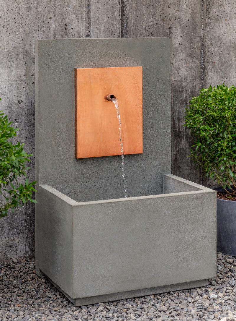 Wall fountain with spout coming out of copper plaque, pictured flanked by two potted shrubs