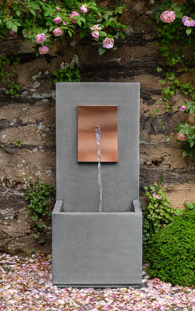 Tall wall fountain with spout coming out of copper plaque pictured in front of stone wall and trailing roses