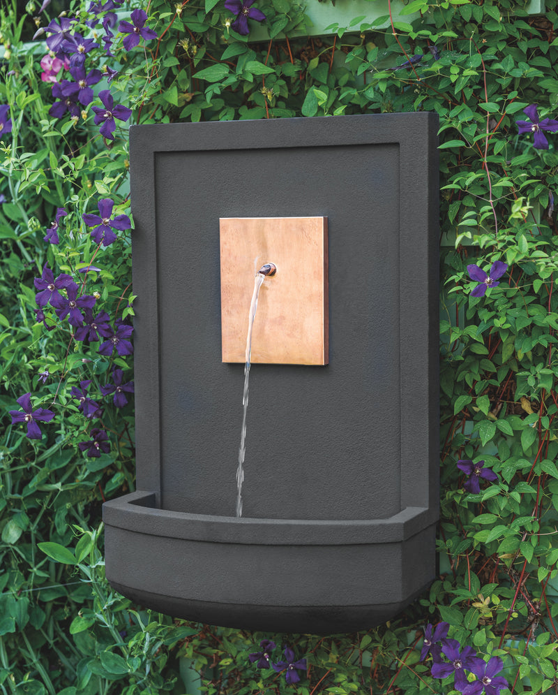 Small black wall fountain with copper plaque shown hanging on wall with clematis