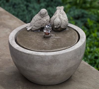 Round tabletop fountain with two birds on the edges and water spouting in front of them pictured on top of bench