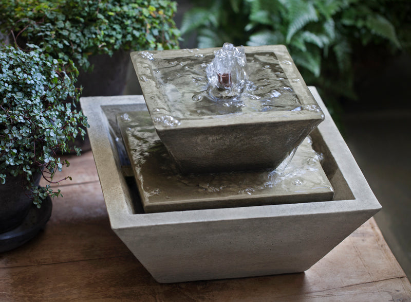 Square tabletop fountain with two levels, pictured on top of table and surrounded by plants