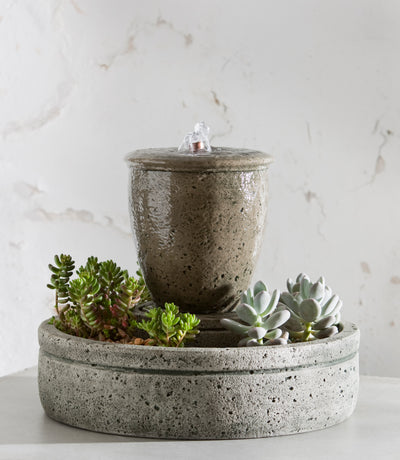 Cylindrical tabletop fountain with a planted basin at the bottom shown in front of white wall