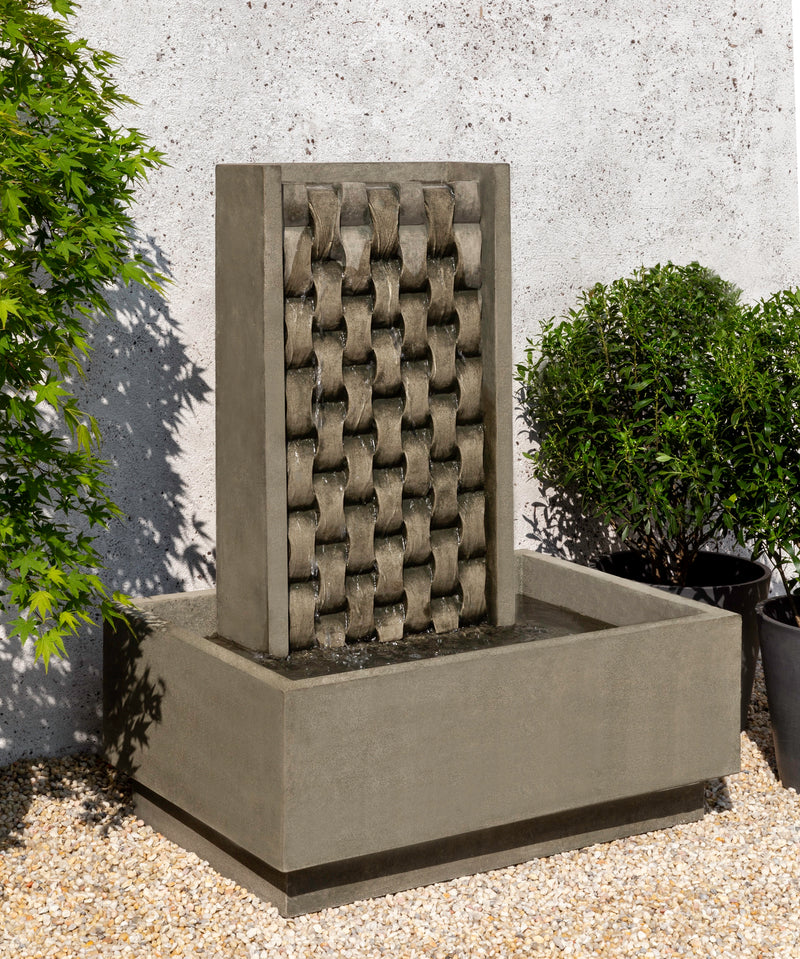 Basin fountain with basket weave design and rectangular basin pictured on gravel in front of wall