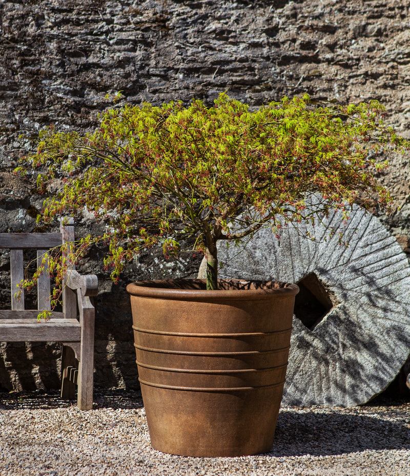 Large brown container planted with a small tree in front of a mill stone and teak bench