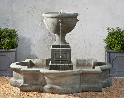 Grey fountain with four spouts running into detailed basin shown in front of white wall