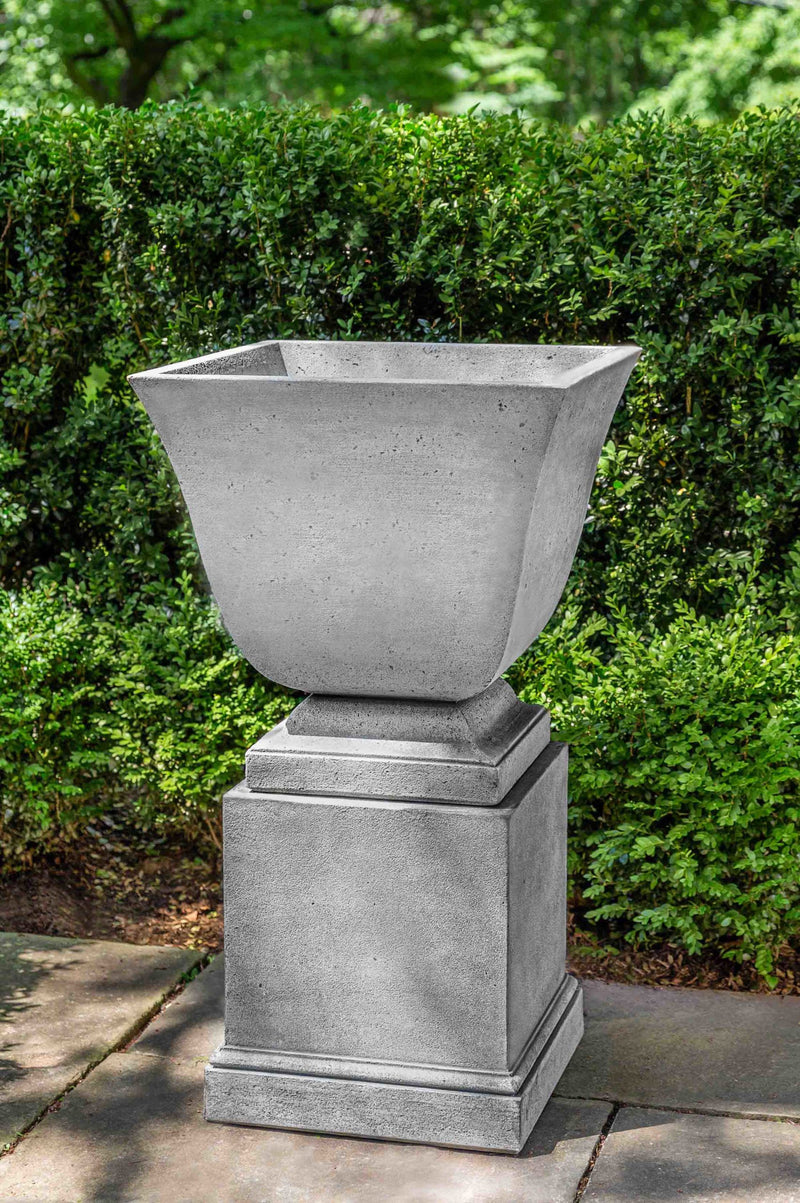 Empty urn shown on top of a pedestal and in front of boxwood hedge