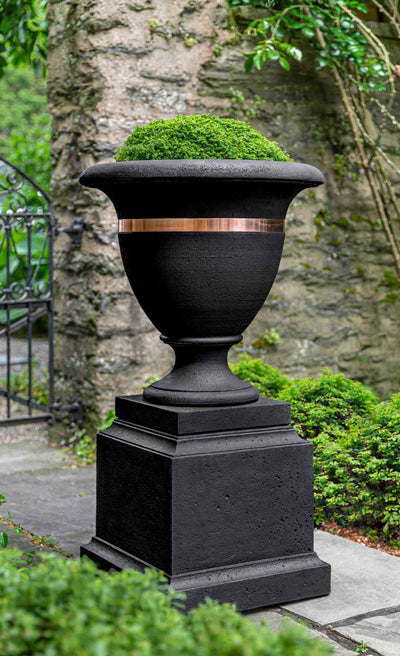 Black urn with a copper band shown on top of a black pedestal in front of a metal gate
