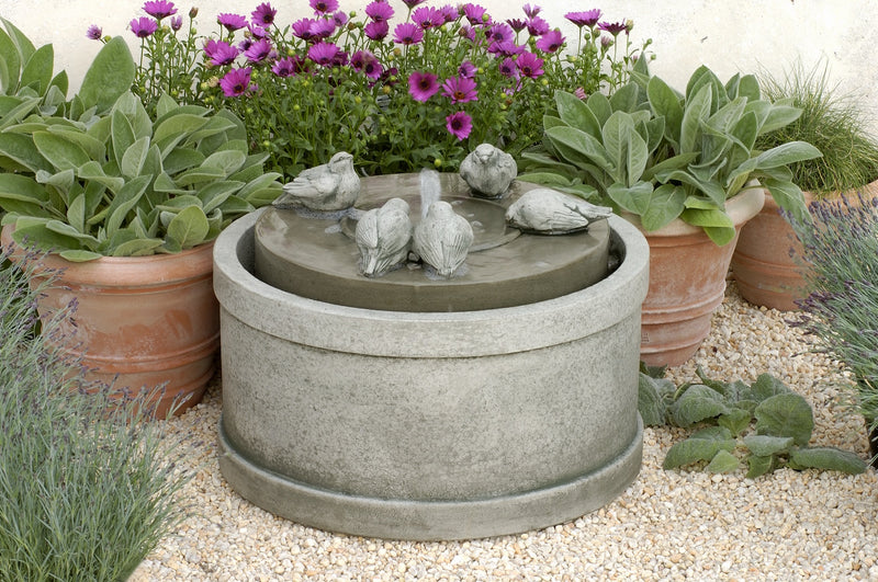 Round fountain with birds sitting on top middle part pictured with potted containers on gravel