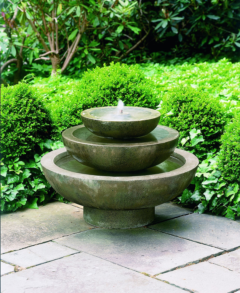 Tiered fountain with three round bowls pictured in front of shrubs