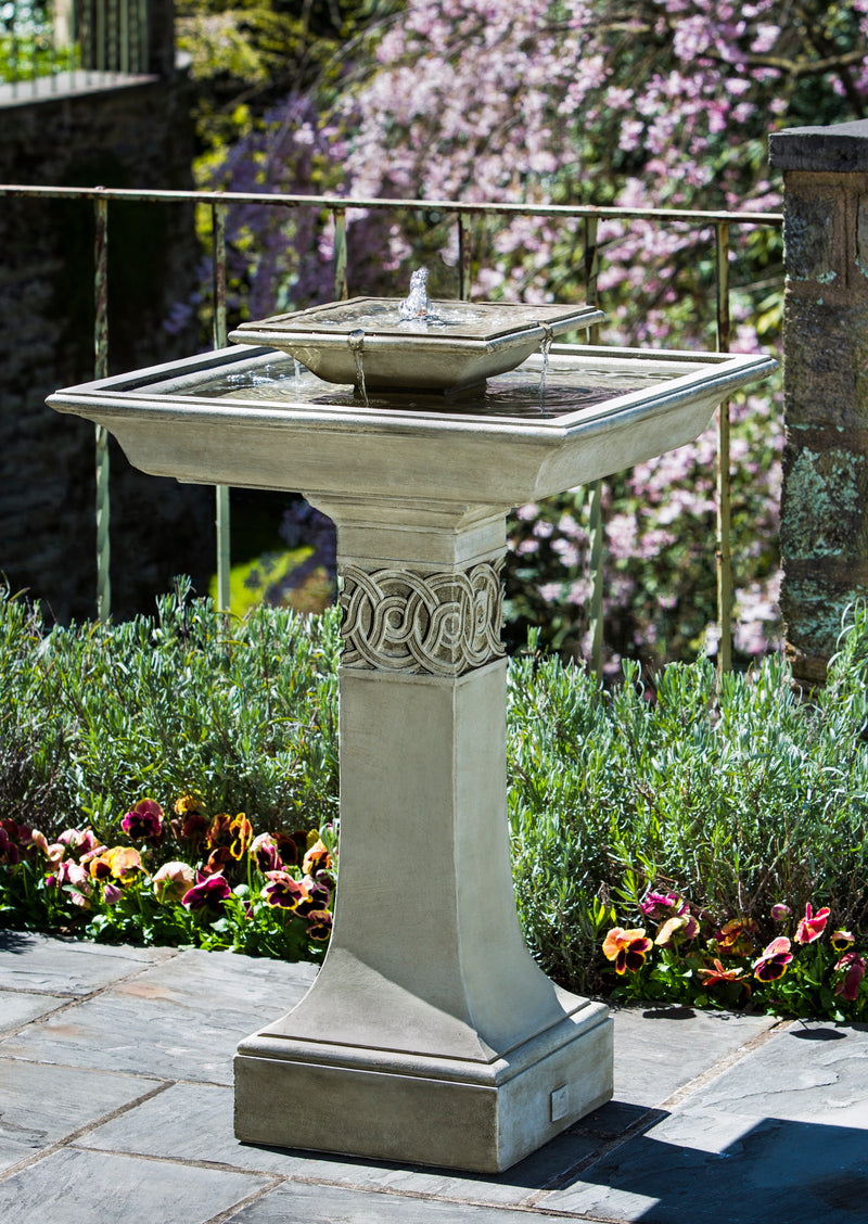 Birdbath fountain with square bowls pictured in front of shrubs