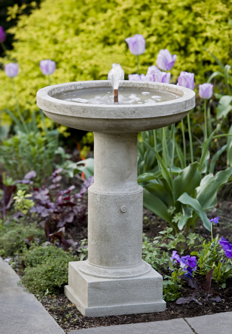 Gray birdbath fountain with medallion on column pictured in front of tulips