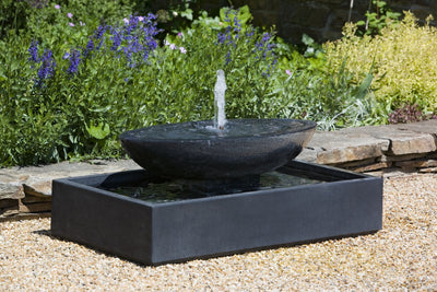 Black fountain with a square basin and oval bowl pictured in front of flower bed