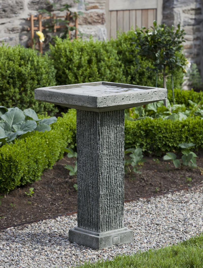 Gray square birdbath with textured pedestal in front of boxwood