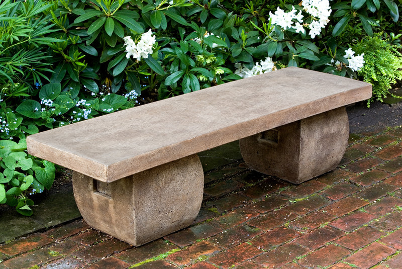 A brown cast stone bench with rounded legs pictured in front of white flowers and on red brick