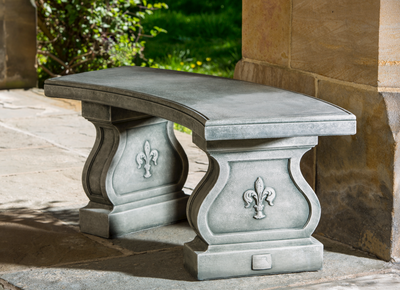 A grey cast stone bench with a curved top and feet decorated with fleur de lys