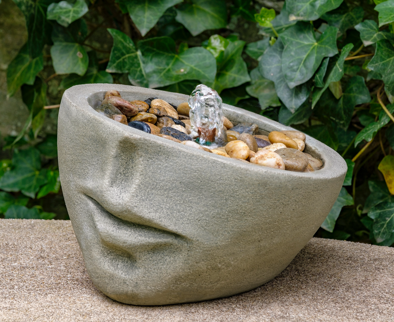 Face shaped table top fountain with pebbles and copper spout pictured  in front of ivy