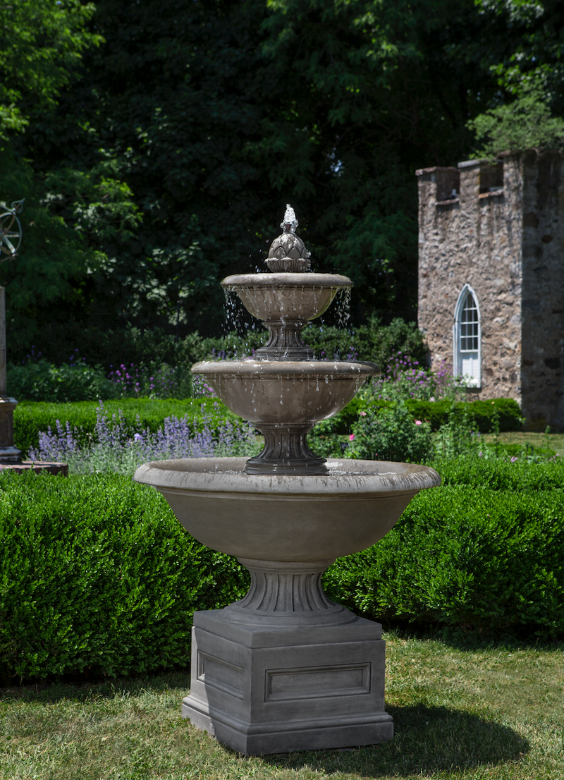 Large three tiered fountain on top of square plinth pictured in front of boxwoods and house in the background