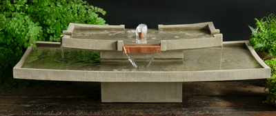Asia-inspired table top fountain with flat copper spout spilling water into lower rectangular bowl, pictured in front of fern