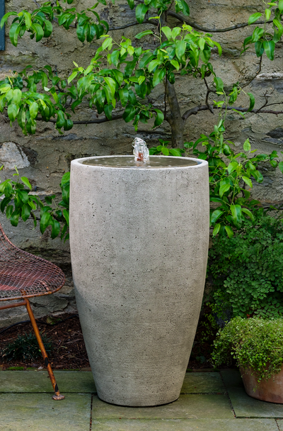 Tall gray fountain with a center spout shown in front of wall and espalier plant