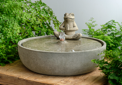 Tabletop fountain with yoga frog sitting on circular top next to ferns