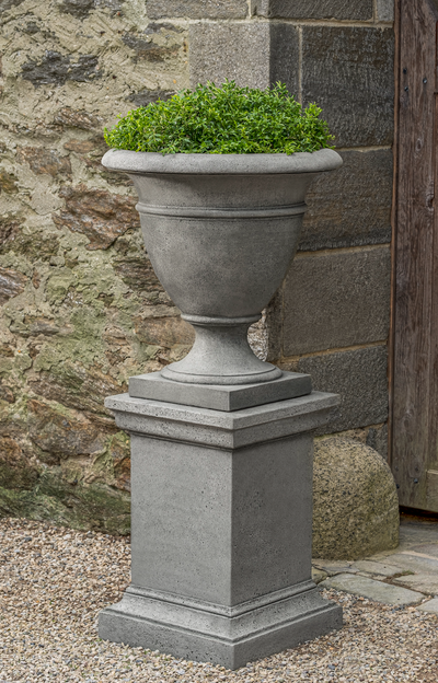 Grey urn on pedestal planted with greenery and placed in front of a stone house
