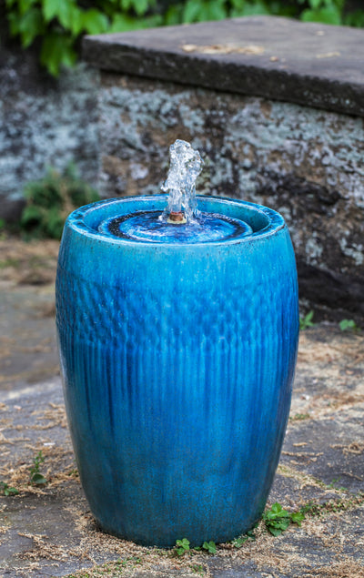 Cylindrical blue glazed fountain with small bubbler pictured in front of stone wall