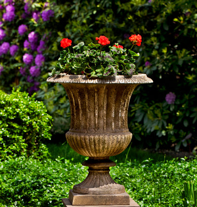 Weathered brown urn planted with red geraniums in front of lush garden