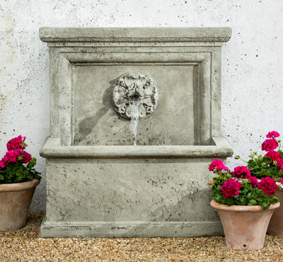 Gray wall fountain with square basin and medallion with copper spout pictured with potted geraniums