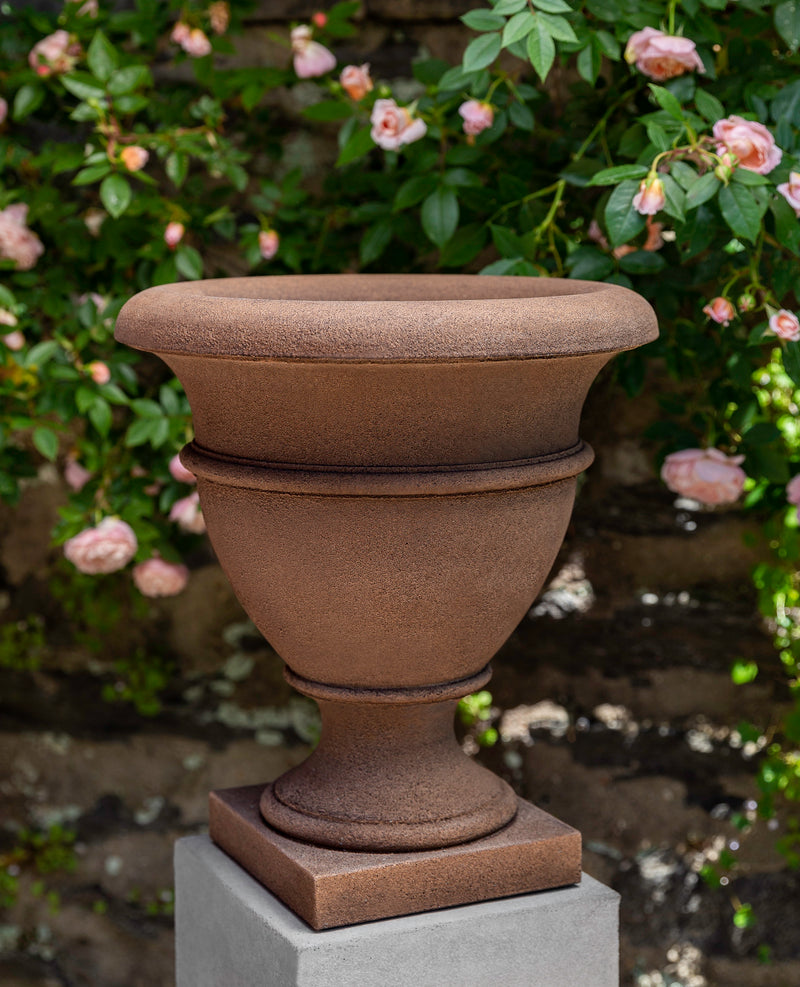 Light brown urn shown in front of pink rose shrubs