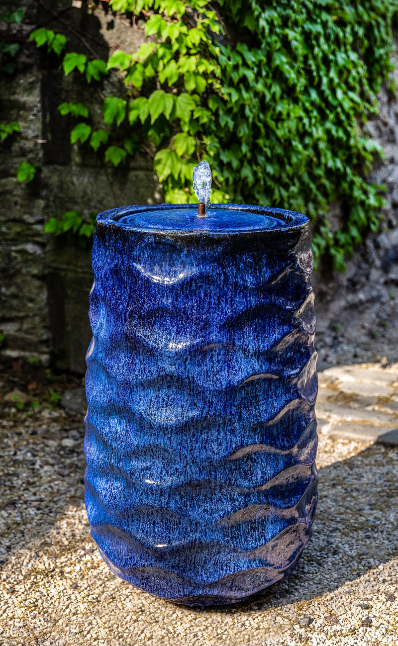 Blue faceted fountain with small bubbler pictured against wall covered in ivy