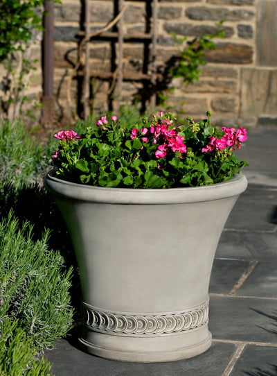 Light grey container with decorative detail at the bottom planted with pink geraniums shown on dark slate floor