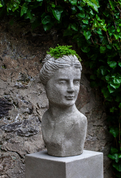 Woman's head shaped container shown on a pedestal in front of a stone wall