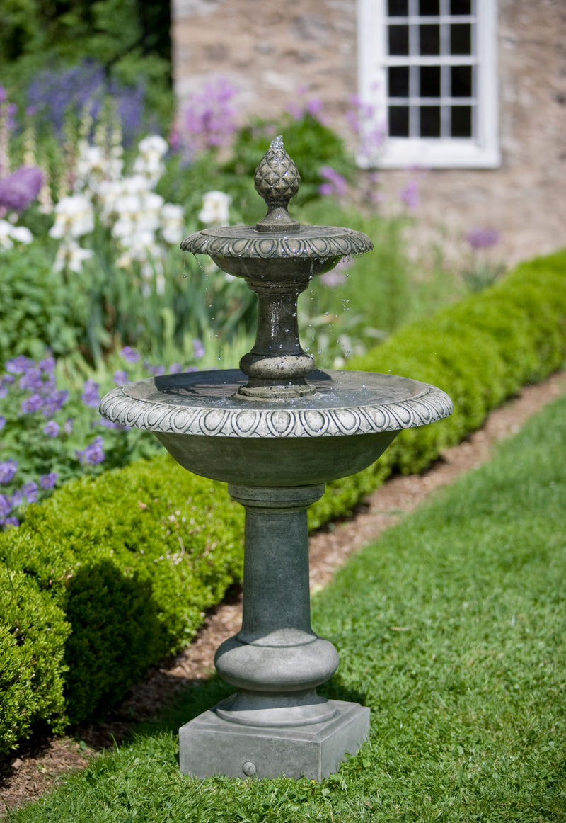 Tiered fountain with pineapple finial pictured in front of shrub edge