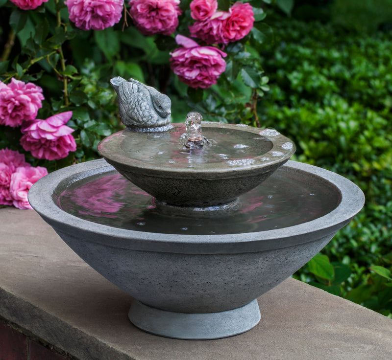 Tabletop fountain with two round bowls and one bird sitting on the top edge pictured in front of roses