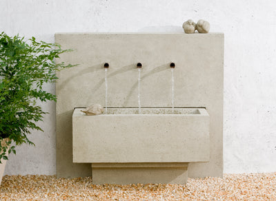 Contemporary wall fountain with three round copper spouts spilling into rectangular basin with two birds sitting on top ledge