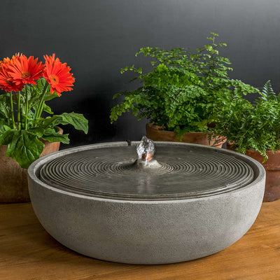 Gray tabletop fountain with rippled top pictured in front of potted plants