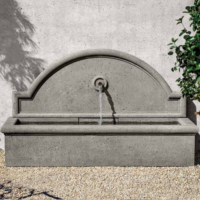A grey fountain with a single spout is against a light grey wall.