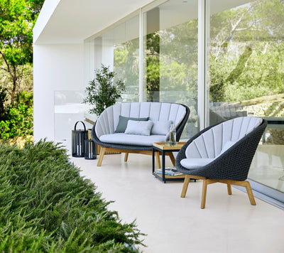 Outdoor sofa and armchair with light grey cushions on narrow patio behind low shrubs