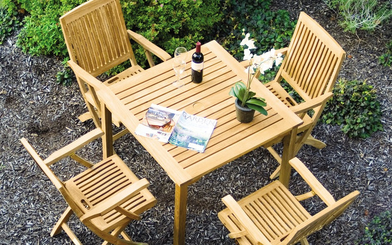 Square teak table with four teak armchairs shown on wood mulch patio